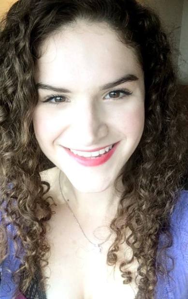 A photo of Lillie Lainoff taken in July 2016. She appears to be white and is wearing a purple shirt and a sea turtle necklace. She has long, curly brown hair and is smiling at the camera.