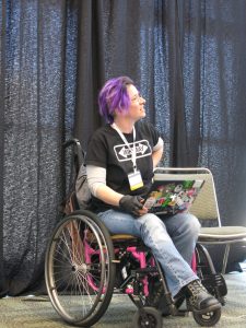 Image description: A woman wearing jeans and a black "Wiki Way" t-shirt, with purple hair, in a pink manual wheelchair with a sticker covered laptop in her lap.