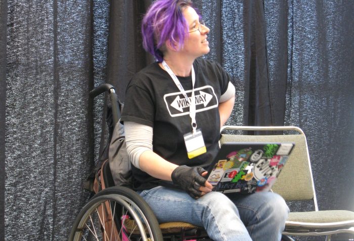 Image description: A woman wearing jeans and a black "Wiki Way" t-shirt, with purple hair, in a pink manual wheelchair with a sticker covered laptop in her lap.