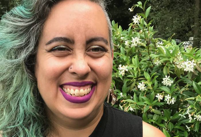 Leah looks at the camera in a garden in South Seattle in August. She has long curly brown, silver and green hair, dark magenta lipstick and light brown skin, and is grinning in front of a garden wall covered in blooming jasmine. Photo credit: Jesse Manuel Graves