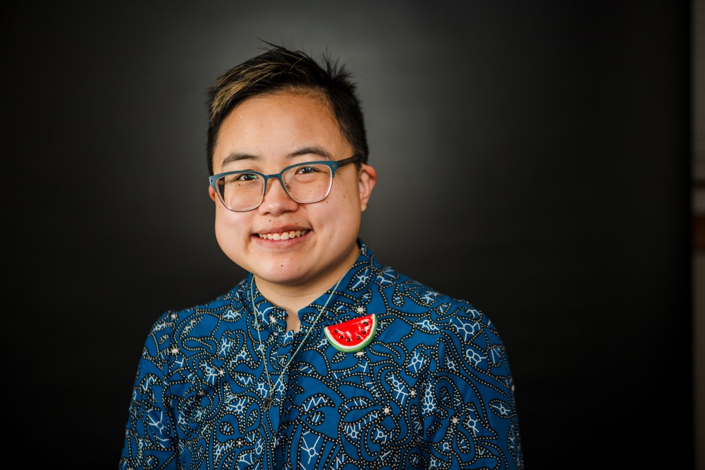 Ly Xīnzhèn is an androgynous and transmasculine East Asian person in their early thirties. They have short black hair with light highlights and they’re wearing a blue top with an abstract pattern, a handmade watermelon pin, and a silver magen david necklace.