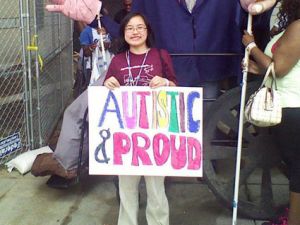 I am standing in front of several people walking down a stone path, wearing khaki pants and a maroon shirt with the text "Organizing autistic people..." visible while holding a large white poster with the colored block letters "Autistic & Proud" in marker. I am a light-complexioned Asian female with short black hair cut slightly past my chin, wearing round glasses.
