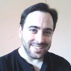 A young white man with dark brown short hair. He has a half-grown beard and is wearing a black shirt. He is smiling at the camera.