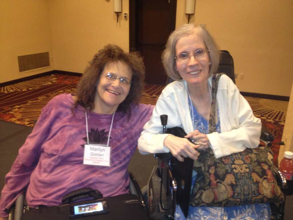 Marilyn Golden [left] in her manual wheelchair with brown curly hair, wire rimmed glasses, purple shirt, conference name tag, a briefcase in her lap, seated next to Diane Coleman [right] in her motorized wheelchair with gray and blond bobbed hair, wire rimmed glasses, blue patterned dress with white sweater, tapestry shoulder bag and slim briefcase to her side. Both are smiling and leaning toward each other. The year is 2014.