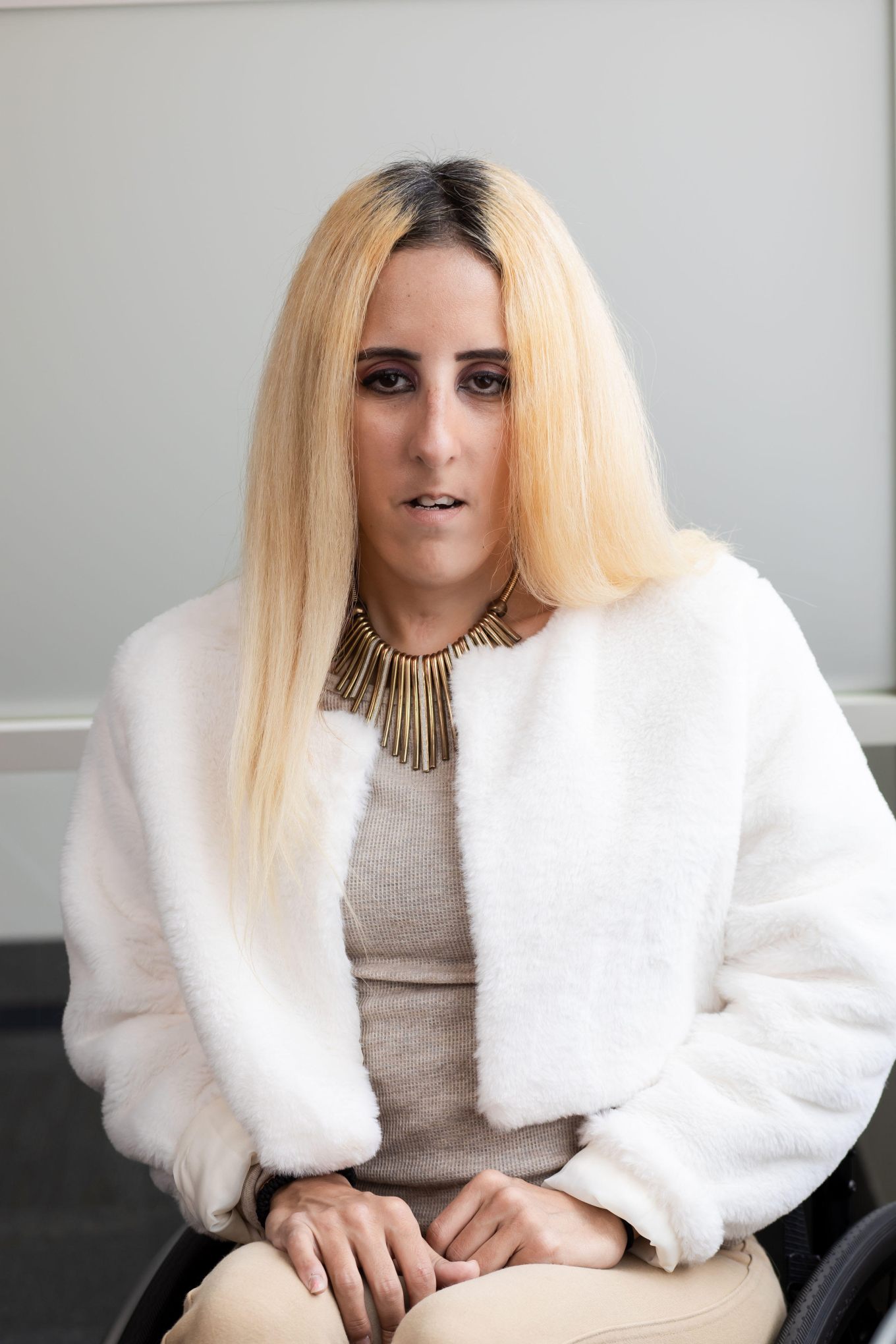 A headshot of a woman with brown color eyes and olive-tan skin complexion with long, straight blonde hair, wearing a baige top with a white jacket and a gold necklace. 