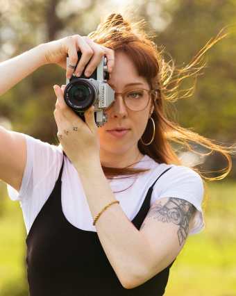 A portrait photo of Marley, a young white woman with red hair, glasses, and tattoos. She stands outdoors and is holding a vintage film camera over her left eye while her right eye is open. The sun illuminates her hair as the wind blows it to the right.
