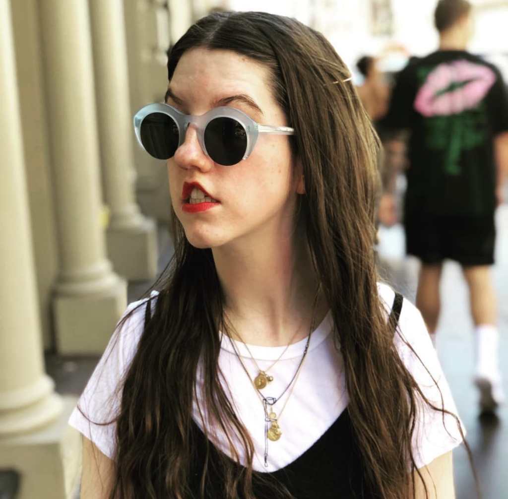 A young, white disabled woman with long dark hair, standing outside and looking off to the side, wearing light blue sunglasses and a white t-shirt with a small black walking stick design and a black sleeveless jumpsuit over the shirt.