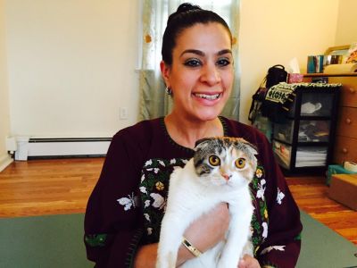 Image Description: Maysoon is smiling. She’s wearing green eyeshadow and her dark hair is in a bun. She is wearing a purple dress with white cross-stitched butterflies and is carrying a Scottish fold calico cat named Beyoncé.