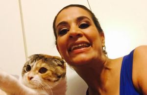 Young Palestinian American woman with her hair pulled back. She is wearing a blue tank top and smiling at the camera. On the left side of the images a Scottish fold cat that is white with streaks of brown and black.