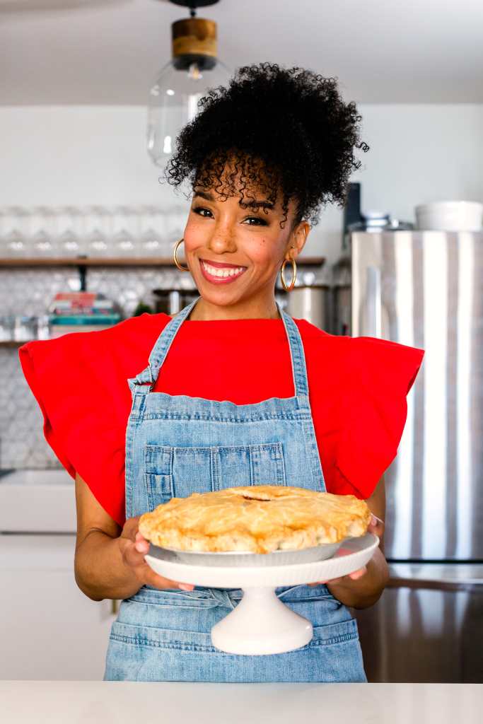 Maya-Camille Broussard, a young Black woman with curly black hair who is wearing gold hoop earrings. She is wearing a bright red top with ruffled short sleeves and a denim apron. She is holding a white ceramic cake stand with a pie that has a golden crust.