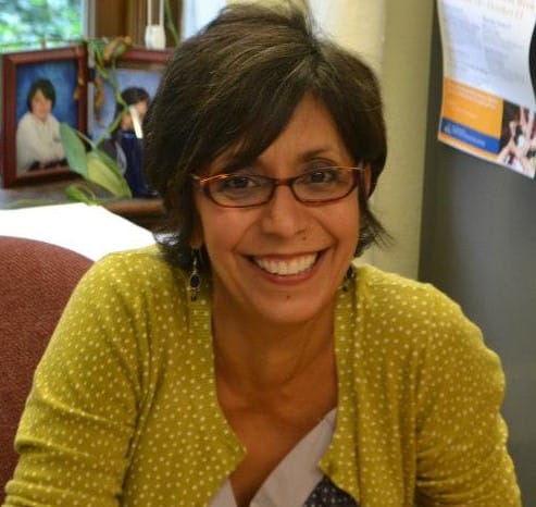 Latina woman with short black and grey hair, wearing brown-rimmed glasses and yellow sweater with white polka dots smiles widely at her desk.