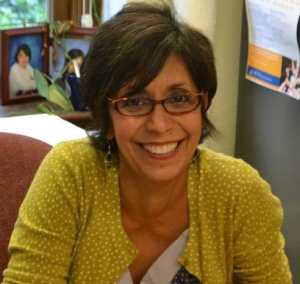Latina woman with short black and grey hair, wearing brown-rimmed glasses and yellow sweater with white polka dots smiles widely at her desk.