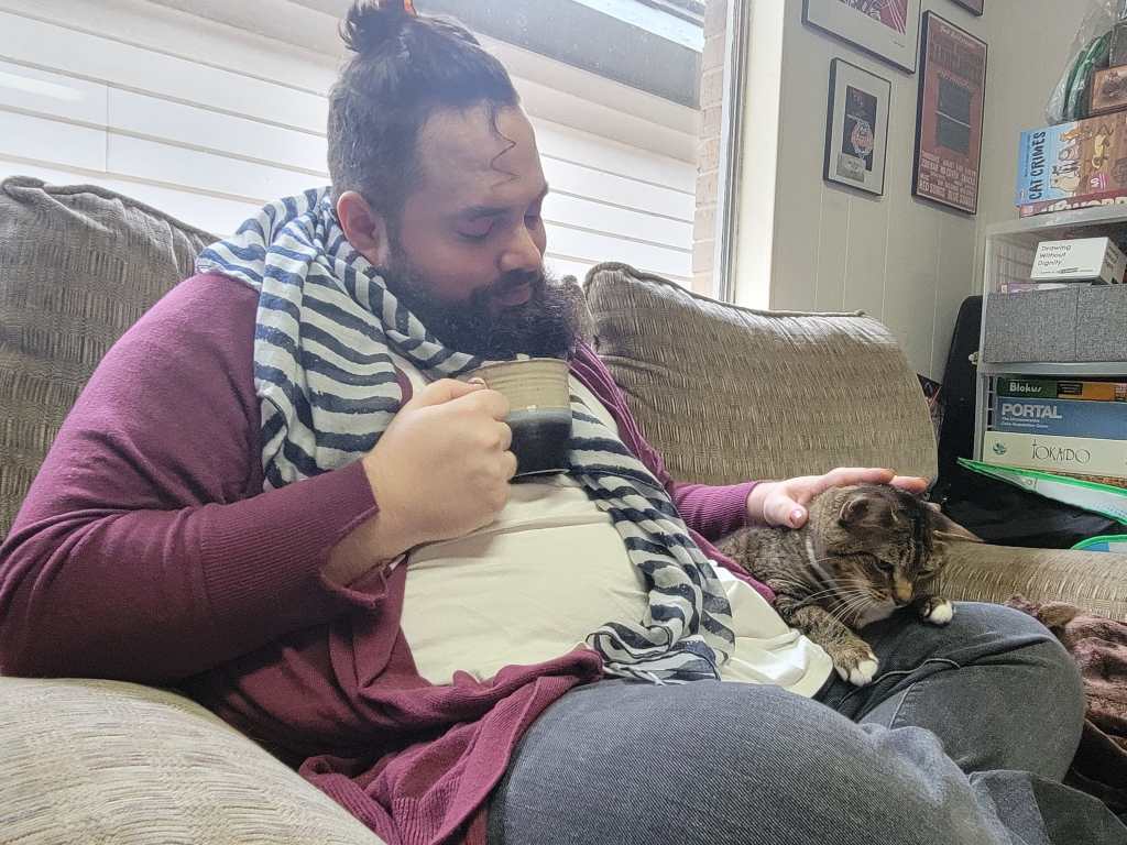 Graphic with a blue-gray background. In the center is a photo of a white, nonbinary, disabled fat person sits on a tan couch with their legs folded. They are wearing a wine red cardigan and a blue striped scarf, and have long, dark curly pulled back in a hair clip and have a dark beard. They are drinking a cup of tea and looking down at a tan cat sitting in their lap and petting the cat.