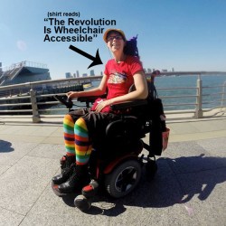 Outdoor photo by the water, a young white woman dressed in rainbow-striped leggings, a red t-shirt and hat is in her wheelchair. She also has a hat and eyeglasses on. In the image is text with a black arrow pointing at her that reads: (shirt reads) 