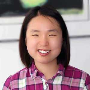 A photo headshot of a young woman with black hair smiling at the camera, wearing a white and plum colored plaid flannel shirt