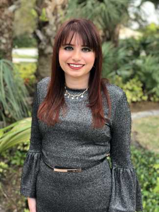 A light-skinned woman with straight red hair and bangs stands, smiling. She has brown eyes, red lipstick, a gold necklace with little moon and star charms on it. Her dress is gray and sparkly with long bell-sleeves and a black and rose-gold belt. Behind her are a ton of green trees 