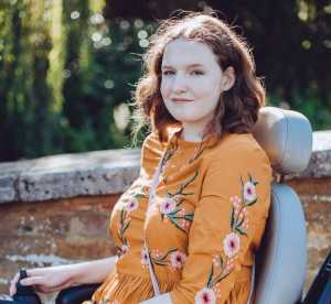 Shona is sitting in her powerchair, facing sideways to the camera, with her head turned to face the camera, she is smiling. She wears a burnt orange coloured top with a floral design on the front of the top and down the arms, which are three quarter sleeves.