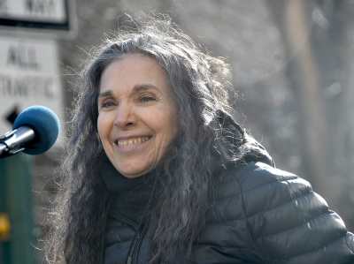 Photo of Nadia LaSpina, an Italian American woman with long curly gray hair with a microphone in front of her. She is wearing a black puffy jacket outdoors.