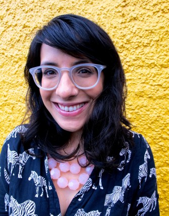 Photo of Neera Jain, a smiling Indian-American woman with wavy shoulder-length brown hair and bangs, clear plastic-framed glasses, and a silver nose ring standing in front of a bright yellow stucco wall. She wears a black shirt-dress printed with white zebras, and a chunky pale pink beaded necklace.