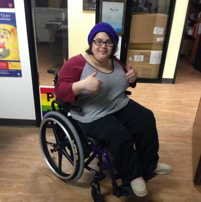 Yolanda Vargas, a young Latinx person in a manual wheelchair wearing a purple knit cap and glasses. She is smiling at the camera with both hands in a thumbs-up position.