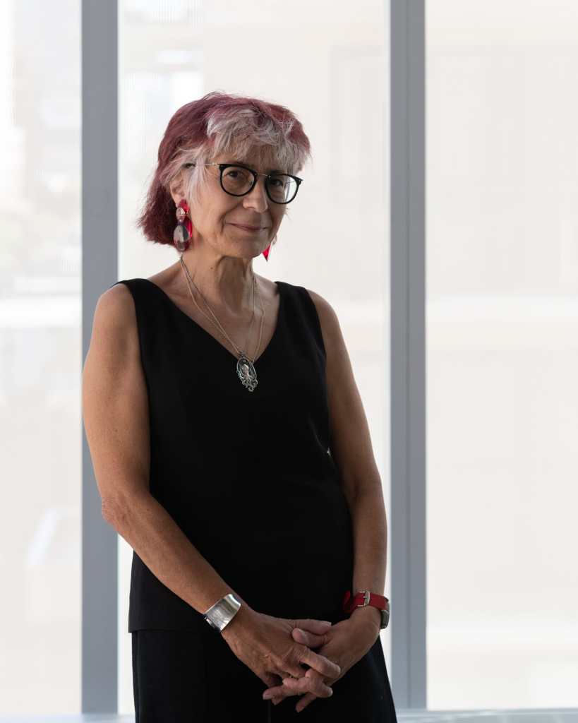 Artist Riva Lehrer, an older white Jewish woman with white hair streaked with red. She is standing against a translucent glass window wearing a sleeveless v-neck black tank and pants. She has a silver necklace, dangly earrings, and glasses on. Her hands are collapsed in front of her torso. Photo credit: Nathan Keay