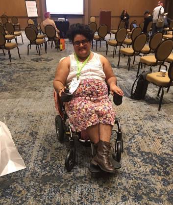 A south asian person poses in a Jazzy Passport folding power chair. He has on brown lace up boots, a colorful floral skirt, and a white lace top. He has a conference badge. He is smiling.