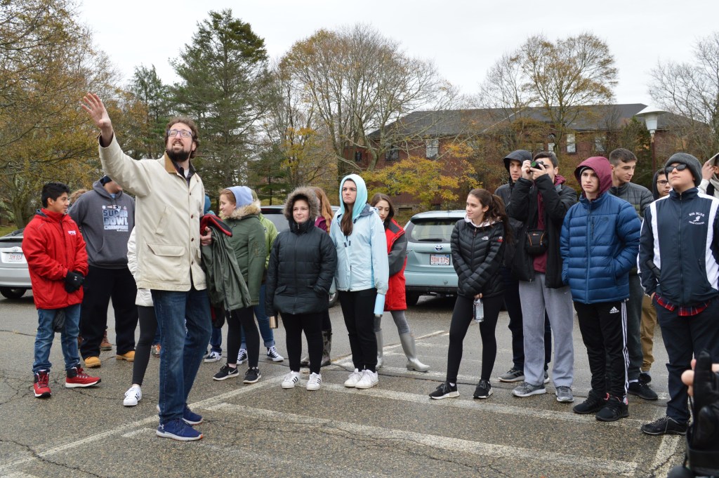 A group of students led by history teacher Alex Green on a field trip to the former Walter E. Fernald Developmental Center. They are standing in a parking lot with Alex pointing toward the Administration Building, which is not pictured. Photo courtesy of Gann Academy. Photo credit: Maya Wainhaus