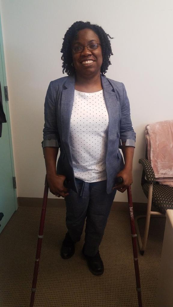 Black woman with curly natural, black hair and glasses in a blue chambray suit and white and blue polka dot top standing on crutches in an office.