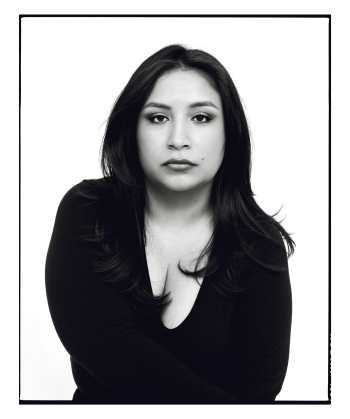Black and white photo of a Latina facing the camera with a serious face, her hair down, and wearing a black v-neck top.