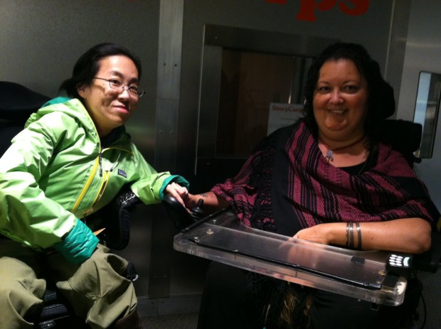 two women of color in wheelchairs in front of a recording booth. The woman on the left is wearing a green jacket, glasses, and has black hair. The woman on the right has long curly dark hair, a black and magenta top and a tray attached to her wheelchair. Both are smiling.