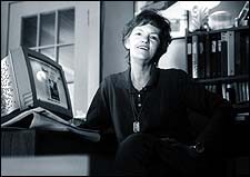 Black and white photo of a white woman with short dark hair. She is wearing a dark shirt and sitting in an office. At the desk is a computer by her side.