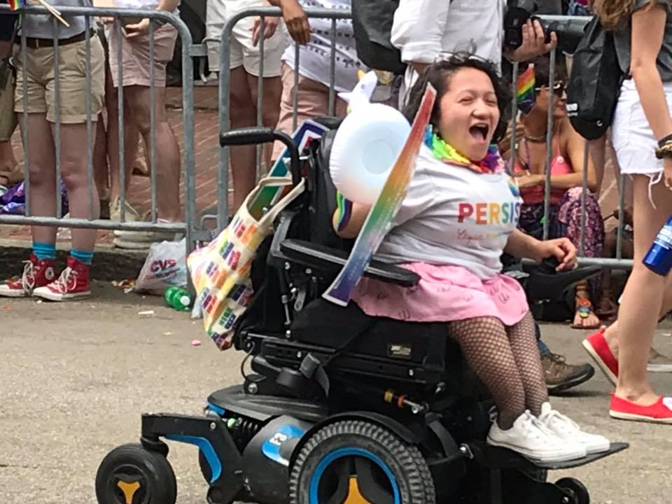 Photo of the author scream yelling at a Boston pride parade in the Elizabeth Warren float. She is seen wearing a rainbow colored PERSIST t-shirt, over a pink dress that has illustrated boobs on it, fishnet tights, and white chuck taylors. She is holding a unicorn blow-up toy and an Elizabeth Warren persist poster. Sandy is sitting in her blue power wheelchair.