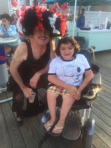 Photo of the author and next to her is one of the iconic Hat Sisters taken in Provincetown at the Boatslip Tea Dance. Sandy is wearing a black and white t-shirt with Brian the Family Guy dog on it that says "Nope, not today" and flannel shorts. She is sitting in a silver power wheelchair. Beside her is Tim O'Conner of the iconic and legendary Hat Sisters. Tim is an older white man wearing a black dress, holding a drink, and an oversized red and white feathered haberdashery fascinator.