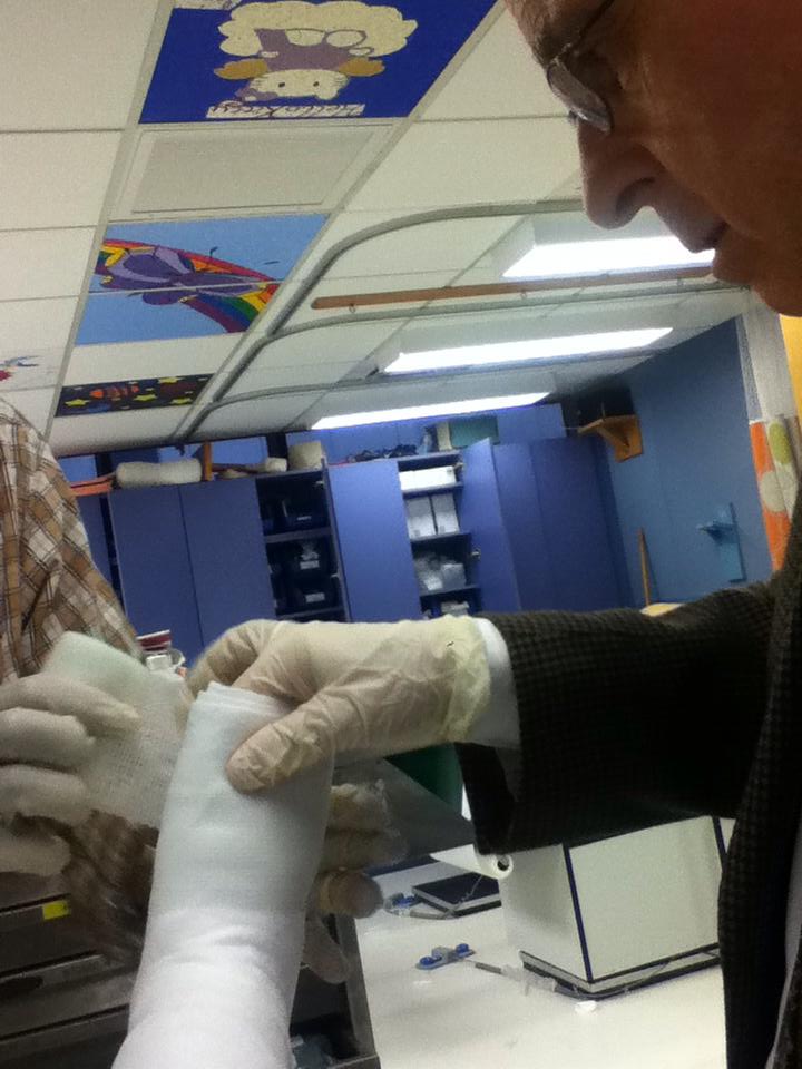 Photo of Dr. Shapiro a white older man wearing glasses, a dark suit jacket, and a gloved hand holding up Sandy's foot that is being wrapped in a white splint. Another person's gloved hand is putting more layers on. The ceiling tiles have illustrations including a cat, butterfly and a rainbow, and a rocket ship with stars. The background is the cast room at Boston Children's Hospital. Blue shelving lines the back walls.