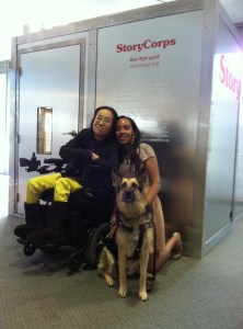 An Asian American woman in a wheelchair next to a young African American woman who knelt down to the same height as the wheelchair user. Between these two people is Maxine, a German shepherd service animal. The three of them are in front of a StoryCorps recording booth.