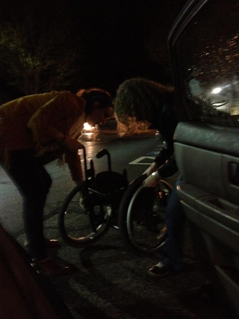 A photo of two of Sandy's friends taking a part her manual wheelchair. Probably after a Starbucks run, or to get ice cream. Raindrops are on the car window, and it is night time. One young woman has shoulder length hair, fringes on her jacket, and is holding onto the handlebar and wheel. The other young woman has curly shoulder length hair, is wearing a leather jacket, a silver bracelet, and she is taking off the other wheel. The photo is taken by the author from inside the passenger seat of the car.