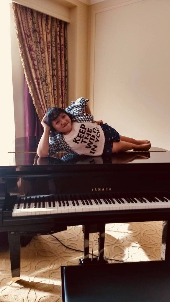 A photo of the author taken by her friend Thomas. She is laying on her side on top of a black Yamaha piano. She has dark shoulder length hair with bangs. Her pink shirt reads Keep The Wild In You, she is also wearing a blue and white flannel shirt. She has dark blue shorts on with rainbows on them. She is holding her head in one hand, and her other arm is on her hip - she is smirking. Red and gold curtains are in the background.