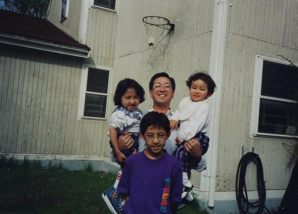 There are 4 people in this photo L-R: a young Sandy with shoulder length hair and wearing a white shirt with blue flowery collar, dark shorts, red tube socks, and denim chuck taylors with the Tasmanian Devil cartoon on them. Her dad, is holder her in one arm and her younger brother in the other. Her dad is smiling proudly, he has dark hair, is wearing large square wire rimmed glasses, and a white collared shirt. Her younger brother has dark curly hair, a white and yellow bib, long white sleeves, plaid pants, and saddle shoes. Her older brother is standing in front with short dark hair, a purple t-shirt with UMBRO lettering on it. In the background is a white house, with a basketball hoop, and guard rails visible in the windows.