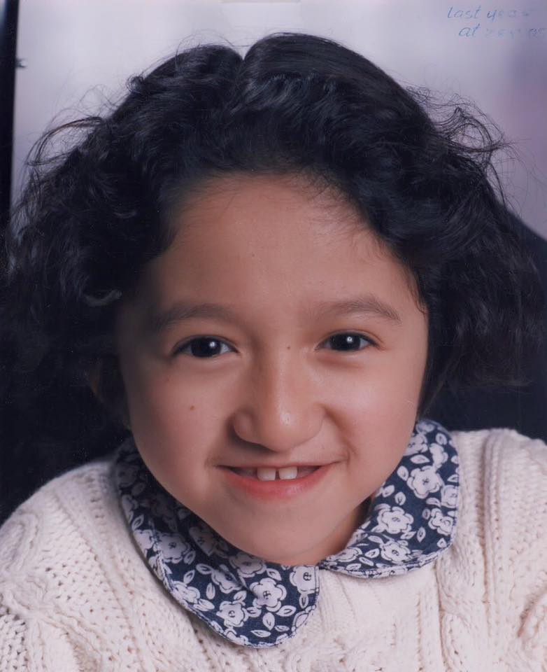 Photo of the author in 5th or 6th grade. She has dark curly hair, is wearing a white sweater, and a blue and white flower collar. She is smiling at the camera.