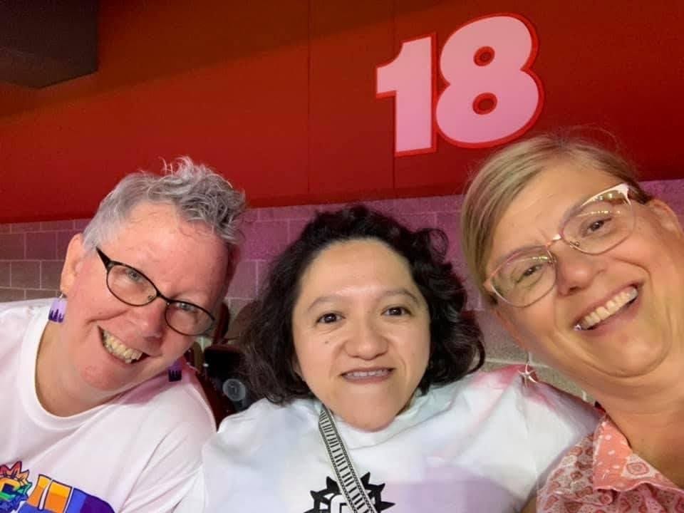 Photo of the author with her dyke moms L-R: Cindy is a white woman with short gray hair, is wearing purple earrings, and dark glasses, she is wearing a white tshirt with rainbow UConn letting on it. Next to her is Sandy, who is wearing a white tshirt with black UConn lettering, and Liz who is a white woman with blonde hair in a pony tail, gold glasses, and is wearing a pink and white collared shirt. They are all smiling at the camera at a UConn women's basketball game.