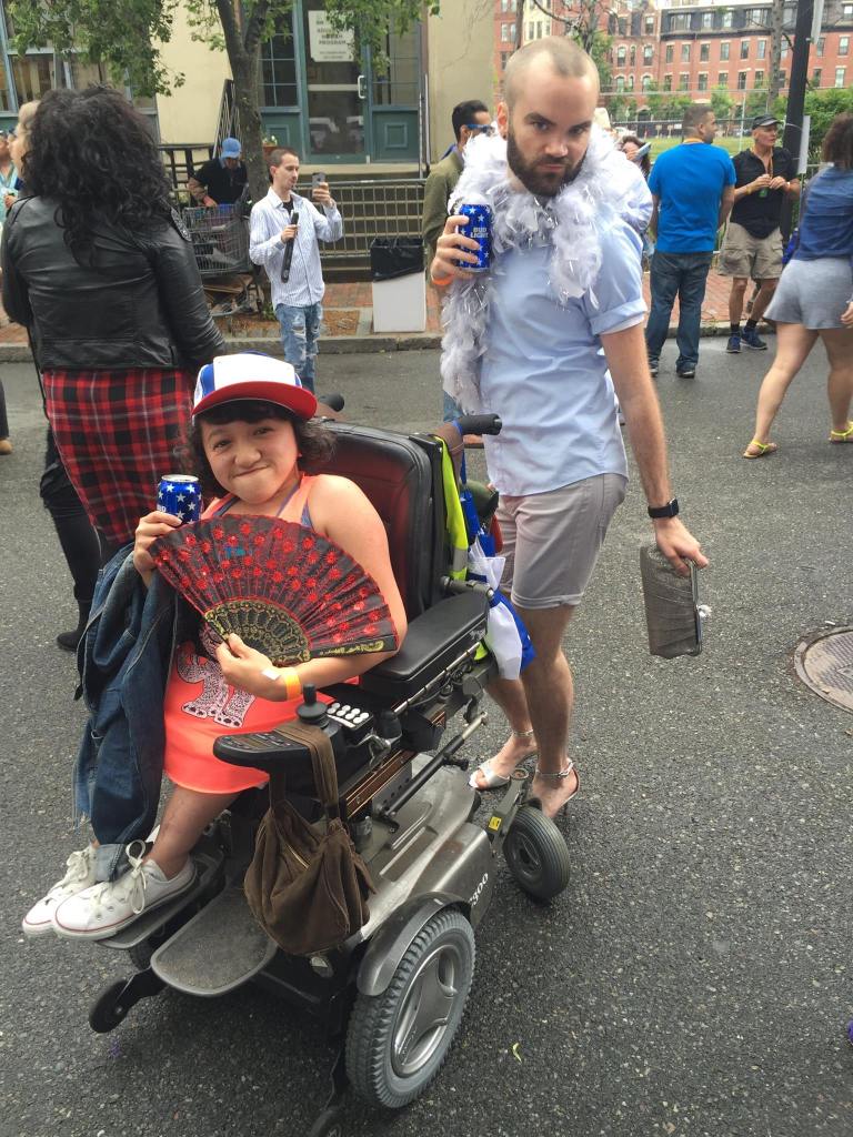 Photo of the author and her friend Thomas outdoors at a Boston pride block party. She is wearing a blue and red snapback hat is holding a decorative fan in one hand, and a drink in the other. She is wearing a neon orange top with a black and white elephant on it, and white chuck taylors. She is sitting in her silver power wheelchair. Thomas is a white man who is posing in silver high heels, a light blue tshirt, and white shorts. He has a white feather boa wrapped around him, in one hand is a silver clutch purse and in the other is a drink.
