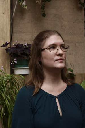 A youngish white woman with chestnut brown hair wearing glasses and a drapey turquoise shirt. She stands in front of a stone wall and potted plants, looking up and away from the camera. Photo credit: Charlie Stern