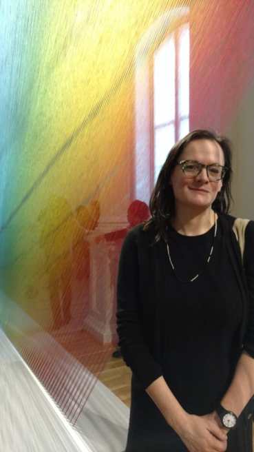Photo of Bess Williamson, a white woman with brown hair and glasses, standing in front of a window and a rainbow thread artwork by Gabriel Dawe.