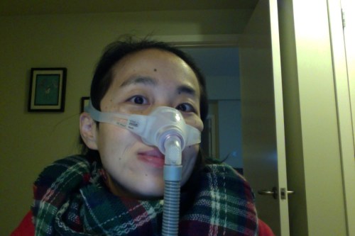 Image of an Asian American woman in her bedroom. She's in a wheelchair and wearing a mask around her nose with a tube. She has a multi-colored plaid scarf around her neck.
