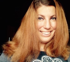 A young white woman against a black background, she has long brown reddish hair. She is smiling at the camera and wearing a gray top with a white lace design on the front