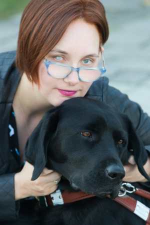 Elsa, a caucasian woman with short asymmetrical red hair, looks over her blue glasses with a half smile on her face. Her right eye is an aqua cataract, her right is grey and has a pupil. She wears pink lipstick that matches her pink and grey hearing aids. She wears a teal geometric blouse and leather jacket, and is pictured with her guide dog. Her guide dog has brown eyes and looks bored.