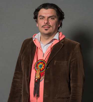 Jean-Luc stands dressed in a button-down shirt, hoodie, and blazer. Hanging from Jean-Luc's neck are a small leather medicine bag and a beaded rosette medallion. Both ornaments are gifts to Jean-Luc from grandmothers in the Mexica tradition.