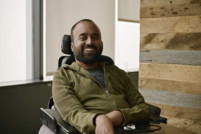Photo of Srin Madipalli, a South Asian man with short black hair and a black beard. He is sitting in a power chair wearing an olive green hoodie. He is smiling at the camera.
