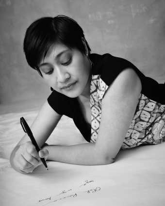 Black and white photo of an Indonesian woman with short hair, earrings, and a patterned dress, lying down on her front, pen in hand, ready to write. Pic credit: Derrick Kakembo.