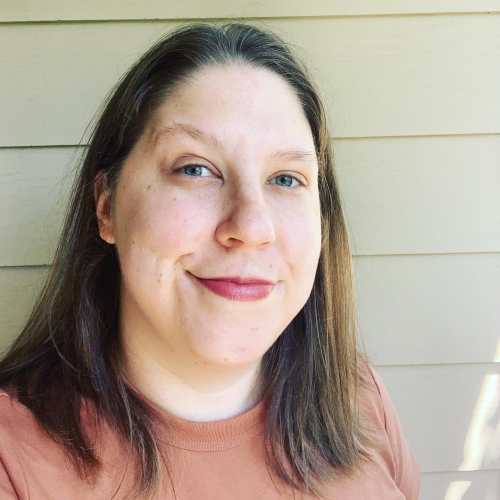 Photo is of Anna, a white nonbinary person with shoulder-length blond hair, standing in front of a beige wall. They wear a muted orange shirt.